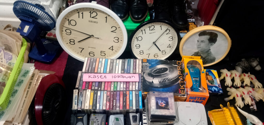 Cluttered display with two large clocks, a framed photo, and a mix of vintage items like cassette tapes and old electronics.