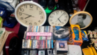 Cluttered display with two large clocks, a framed photo, and a mix of vintage items like cassette tapes and old electronics.
