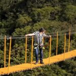 People cross a bright orange suspension bridge over a forest; man in striped shirt wearing a harness, woman at far end waiting.