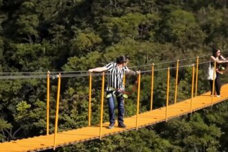 People cross a bright orange suspension bridge over a forest; man in striped shirt wearing a harness, woman at far end waiting.