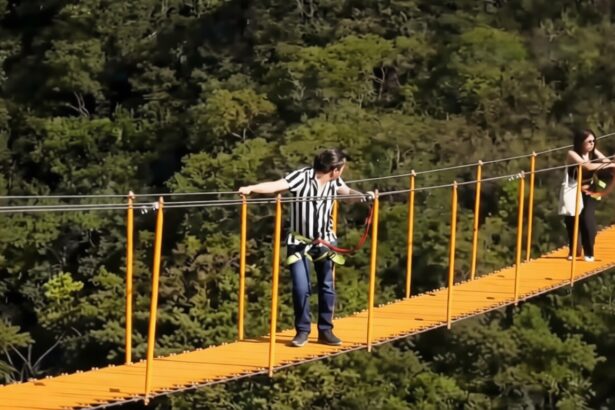 People cross a bright orange suspension bridge over a forest; man in striped shirt wearing a harness, woman at far end waiting.