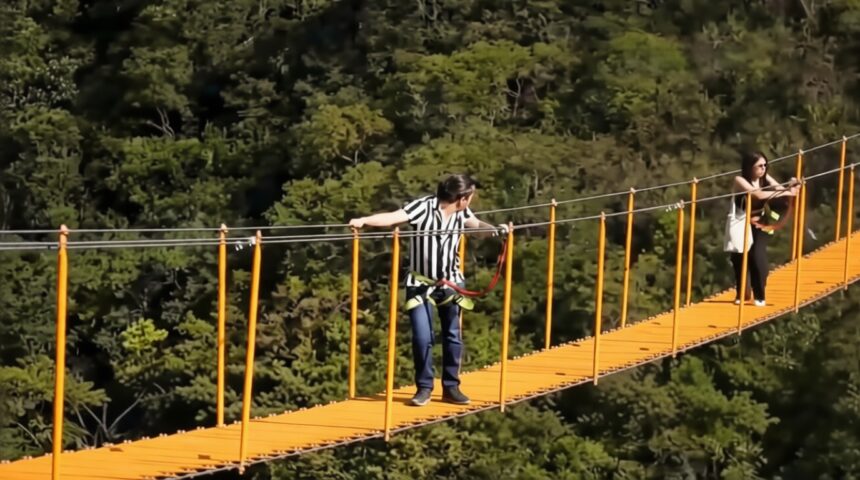People cross a bright orange suspension bridge over a forest; man in striped shirt wearing a harness, woman at far end waiting.