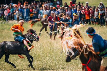 Riders on galloping horses compete in a traditional grassy-field race, with a large crowd watching in the background.