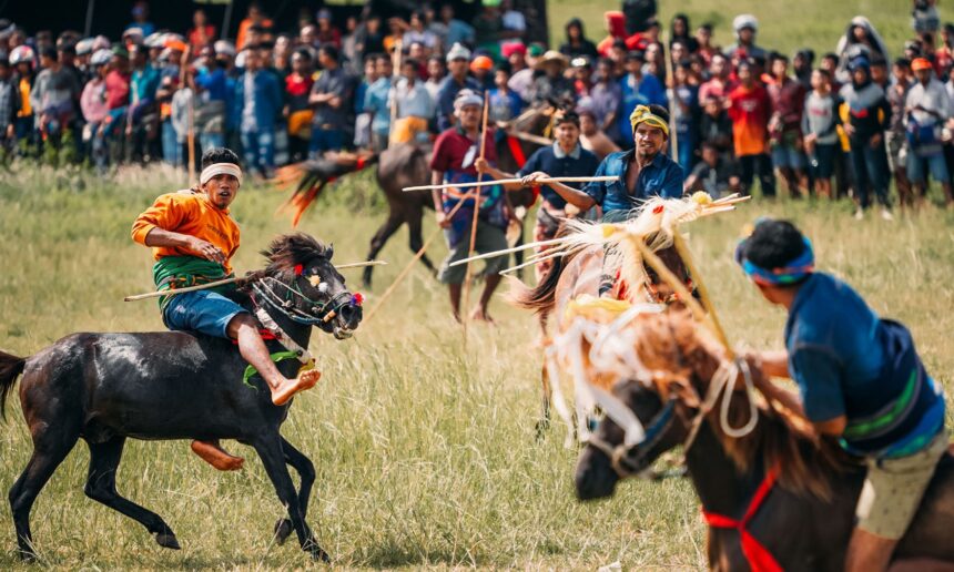 Riders on galloping horses compete in a traditional grassy-field race, with a large crowd watching in the background.