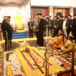 Royal ceremony around a throne; officials in black and gold stand in a grand hall while children in yellow sit on the floor bedded by a colorful rug.