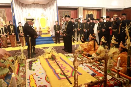 Royal ceremony around a throne; officials in black and gold stand in a grand hall while children in yellow sit on the floor bedded by a colorful rug.