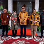 Group of five people standing on a red carpet in a modern building lobby, wearing batik outfits for a formal photo.