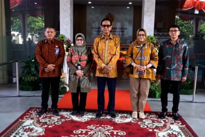 Group of five people standing on a red carpet in a modern building lobby, wearing batik outfits for a formal photo.