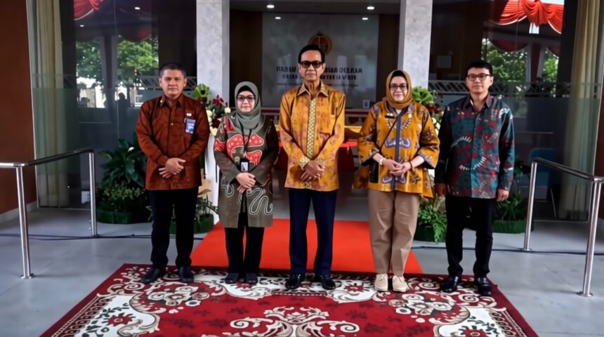 Group of five people standing on a red carpet in a modern building lobby, wearing batik outfits for a formal photo.