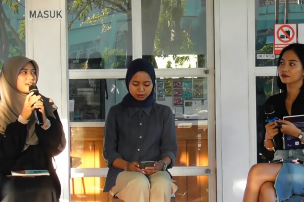 Three women sit on a stage in a glass-walled room: left speaks into a microphone, middle woman in a hijab looks at her phone, right holds a notebook.