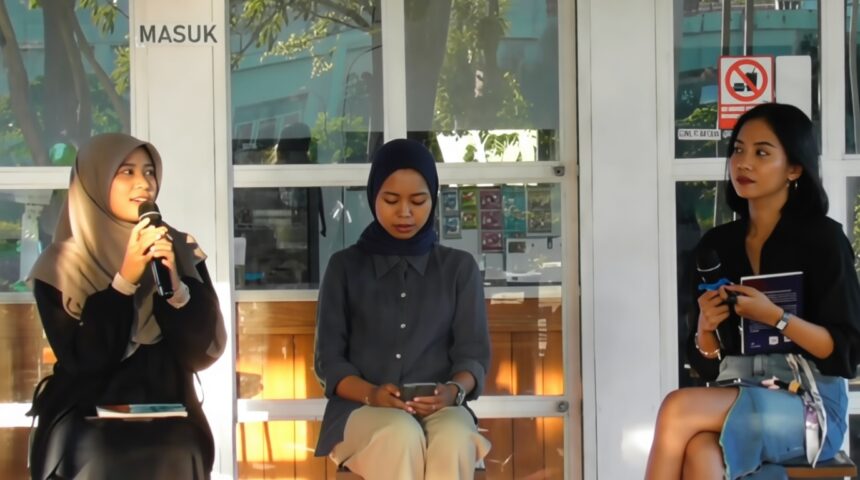 Three women sit on a stage in a glass-walled room: left speaks into a microphone, middle woman in a hijab looks at her phone, right holds a notebook.