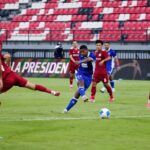 Soccer scene: a blue-team player strikes the ball as red defenders close in on a green field with red-and-white stadium seating in the background.