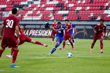 Soccer scene: a blue-team player strikes the ball as red defenders close in on a green field with red-and-white stadium seating in the background.