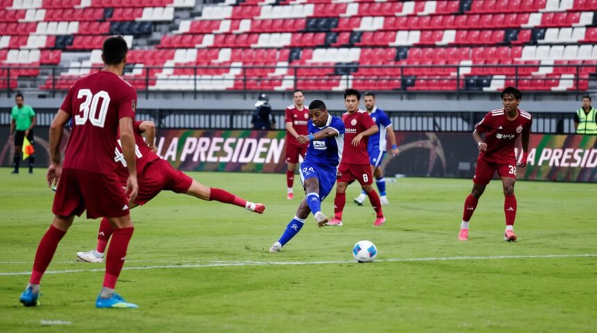 Soccer scene: a blue-team player strikes the ball as red defenders close in on a green field with red-and-white stadium seating in the background.