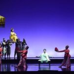 Stage performance: dancers in red surround a pale-green-clad performer center stage; a yellow-dressed figure sits elevated on a platform held by others under a parasol.