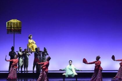 Stage performance: dancers in red surround a pale-green-clad performer center stage; a yellow-dressed figure sits elevated on a platform held by others under a parasol.