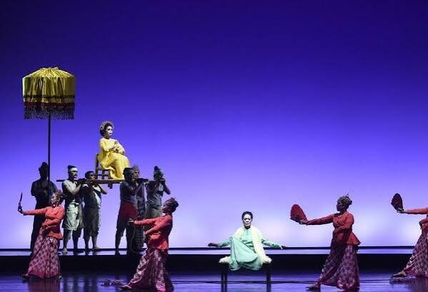 Stage performance: dancers in red surround a pale-green-clad performer center stage; a yellow-dressed figure sits elevated on a platform held by others under a parasol.