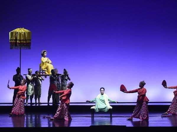 Stage performance: dancers in red surround a pale-green-clad performer center stage; a yellow-dressed figure sits elevated on a platform held by others under a parasol.