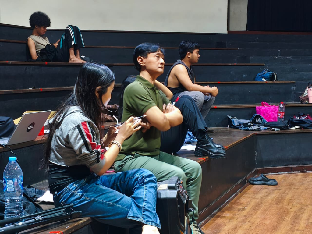 People sit on dark wooden bleachers in a rehearsal space; a woman with a laptop and a man in a green polo rest in the foreground.
