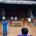 People rehearsing on a stage with chairs, a table, and a ladder in the background, under dark curtains and stage lights on a wooden floor.