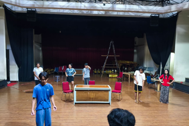 People rehearsing on a stage with chairs, a table, and a ladder in the background, under dark curtains and stage lights on a wooden floor.