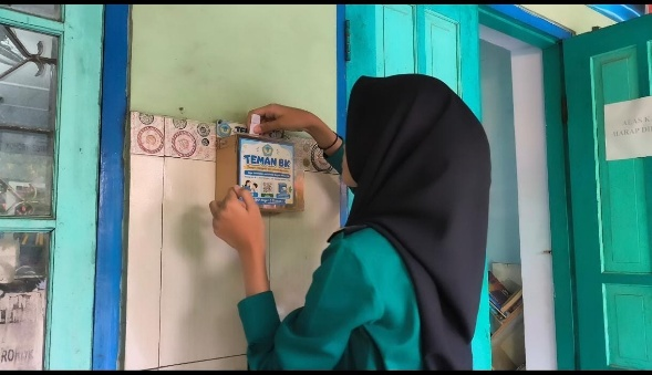 A woman in teal with a black hijab attaches a small blue-and-cream TEMAN BK box to a tiled wall beside a turquoise door.