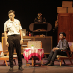 Stage scene with two men: a uniformed officer stands while a man in a dark shirt sits in a chair, a small round table with a pink tablecloth between them and a cluttered cabinet backdrop.