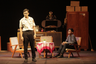 Stage scene with two men: a uniformed officer stands while a man in a dark shirt sits in a chair, a small round table with a pink tablecloth between them and a cluttered cabinet backdrop.