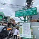 Street sign at an urban intersection reading 'Jl. Soedarna Tresna Manggala' with additional local script beneath, mounted on a green post above a busy street. Nearby buildings and vehicles are visible in the background.