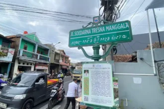 Street sign at an urban intersection reading 'Jl. Soedarna Tresna Manggala' with additional local script beneath, mounted on a green post above a busy street. Nearby buildings and vehicles are visible in the background.