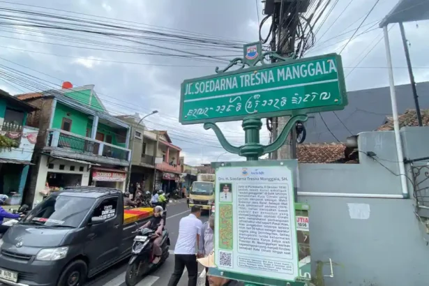 Street sign at an urban intersection reading 'Jl. Soedarna Tresna Manggala' with additional local script beneath, mounted on a green post above a busy street. Nearby buildings and vehicles are visible in the background.