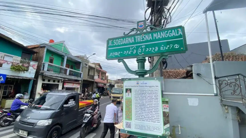 Street sign at an urban intersection reading 'Jl. Soedarna Tresna Manggala' with additional local script beneath, mounted on a green post above a busy street. Nearby buildings and vehicles are visible in the background.