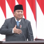 Man in a dark suit and black peci hat speaks at a podium with two microphones, red-and-white flags behind him.