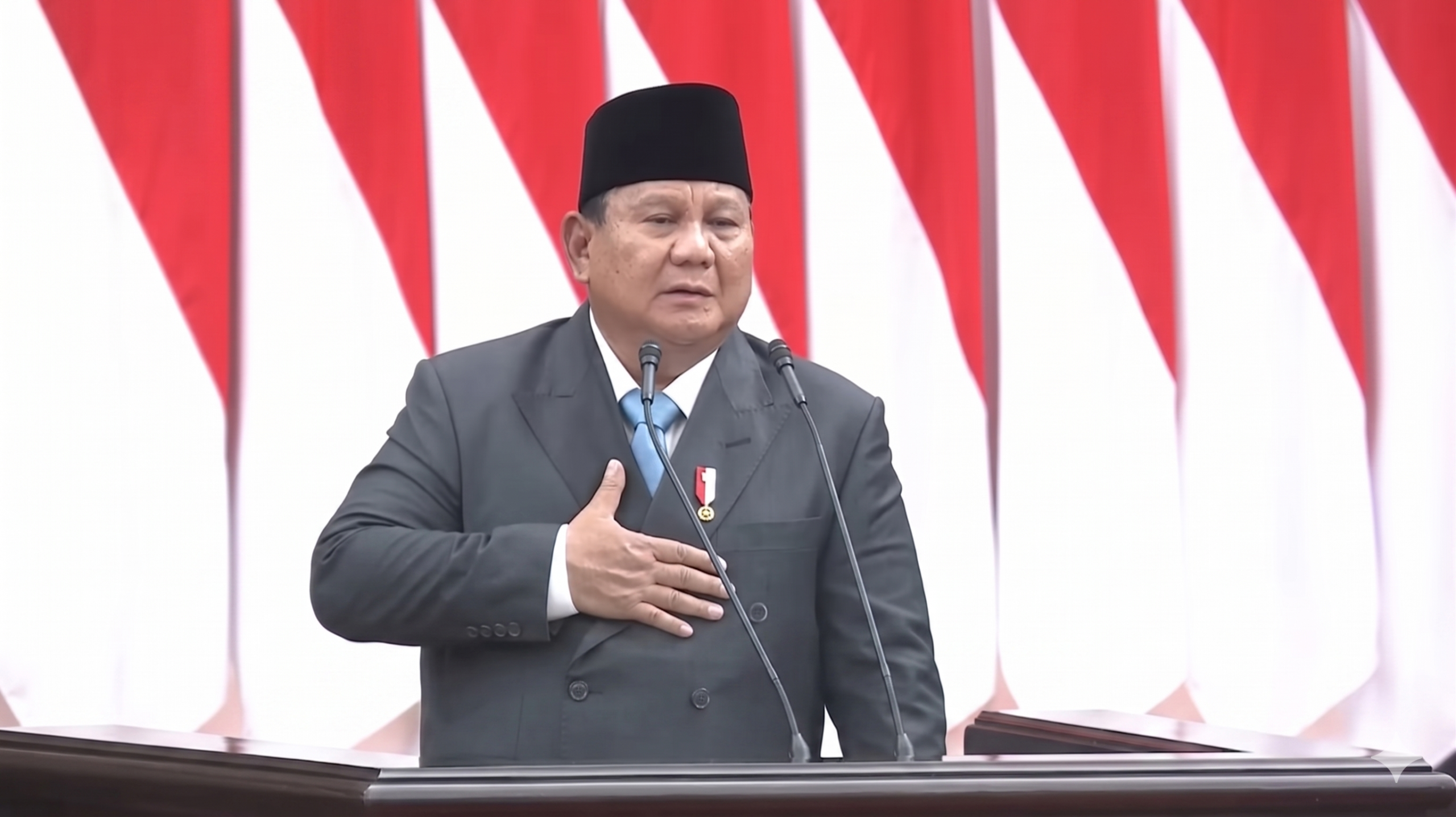 Man in a dark suit and black peci hat speaks at a podium with two microphones, red-and-white flags behind him.