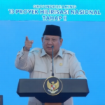 Government official delivering a speech at a podium during a national project groundbreaking event, blue backdrop with Indonesian emblems and signage