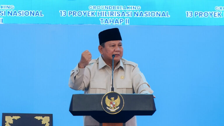 Indonesian official in beige uniform and black cap speaks at a podium with national emblem, gesturing with a raised fist during a groundbreaking ceremony backdrop reading '13 Proyek Hilirisasi Nasional Tahap II'