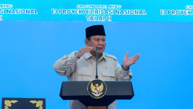 Indonesian official in beige uniform and peci speaks at a podium with the national emblem; blue backdrop reads '13 Proyek Hilirisasi Nasional Tahap II'.