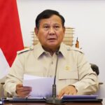 A man in a beige uniform seated at a desk with papers, speaking into a microphone during a formal briefing. A red-and-white flag is in the background.