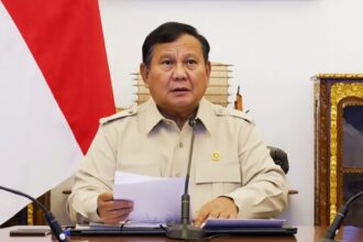 A man in a beige uniform seated at a desk with papers, speaking into a microphone during a formal briefing. A red-and-white flag is in the background.