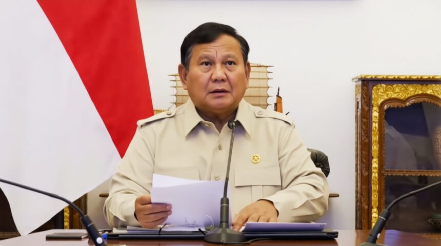 A man in a beige uniform seated at a desk with papers, speaking into a microphone during a formal briefing. A red-and-white flag is in the background.