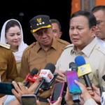 Man in beige uniform speaks to reporters as microphones surround him outside a building