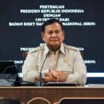 Indonesian official at a press conference, seated at a desk with a small flag and microphone, backdrop with white text in Indonesian