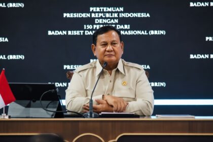 Indonesian official at a press conference, seated at a desk with a small flag and microphone, backdrop with white text in Indonesian