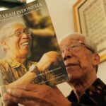 Older man in a batik shirt holding a magazine titled 'Sejarah Indonesia: Perspektif Lokal dan Global,' featuring a laughing elderly woman on the cover.