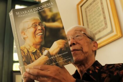 Older man in a batik shirt holding a magazine titled 'Sejarah Indonesia: Perspektif Lokal dan Global,' featuring a laughing elderly woman on the cover.