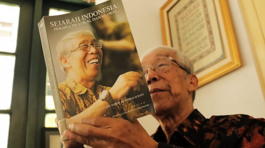 Older man in a batik shirt holding a magazine titled 'Sejarah Indonesia: Perspektif Lokal dan Global,' featuring a laughing elderly woman on the cover.