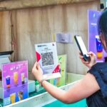Vendor wearing a mask at a colorful drink stall, assisting a customer who holds a QR menu flyer.