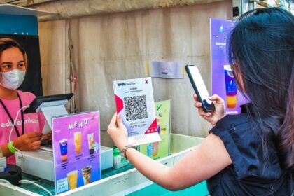 Vendor wearing a mask at a colorful drink stall, assisting a customer who holds a QR menu flyer.