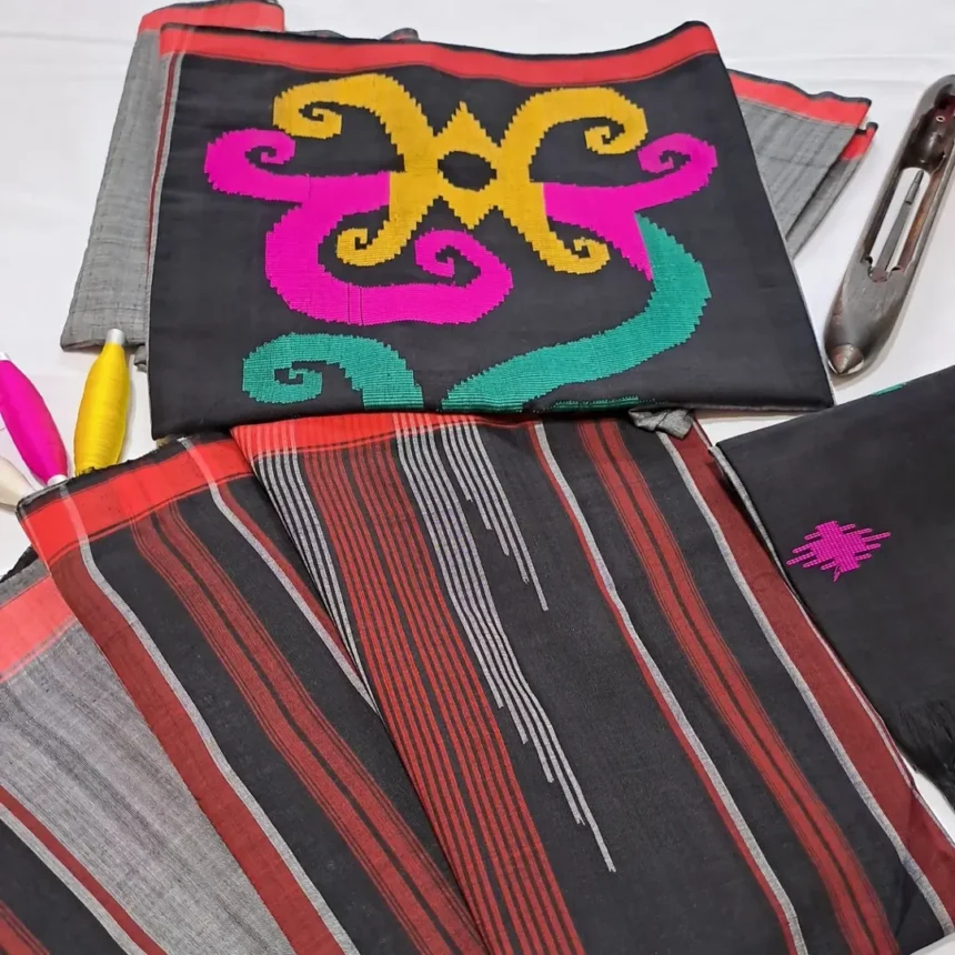 RTR - Kain Sarung Samarinda - Sarung Tenun ATBM - SARIMBIT Motif pakis dan Motif pucuk bambu - Mabur.co Black fabric with bright yellow and pink embroidery, placed on striped red-gray textiles with sewing tools nearby.