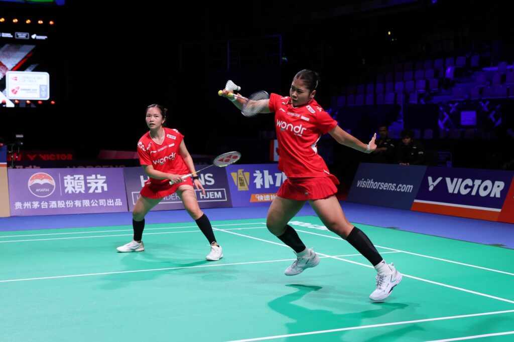 Two female badminton players in red uniforms compete on a bright green court, one reaching with her racket for a shot, shuttlecock in play.